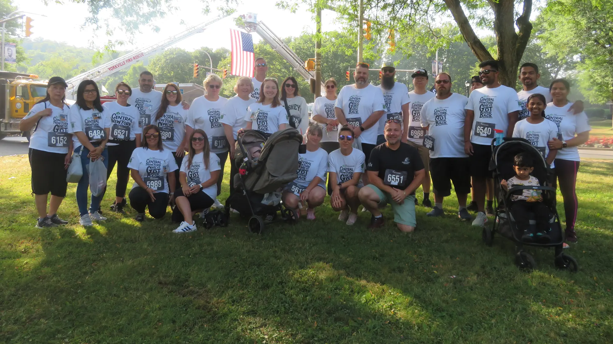 The Fountainhead Group team wearing race bibs and Central New York We&rsquo;ve Got Your Back shirts, gathered under a tree beside a fire department ladder truck flying the American flag